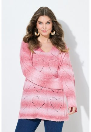 Kvinde med bølget brunt hår iført store gyldne øreringe og en lyserød strikket sweater med hjerte mønstre, kombineret med blå jeans, mod en hvid baggrund.