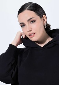 Jonge vrouw met kort, donker haar en oorbellen met een ring, gekleed in een zwarte hoodie, die de kraag van de hoodie aanraakt, tegen een effen achtergrond.