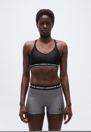 Persona atlética de pie mirando hacia adelante, vestida con un sujetador deportivo negro de Under Armour y pantalones cortos ajustados grises de Under Armour, sobre un fondo liso.