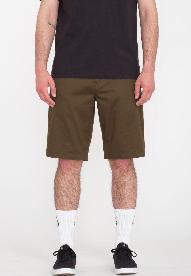 Olivgrüne Shorts mit einer lockeren Passform, knielangem Saum, Fronttaschen und einem glatten Baumwoll-Mix-Stoff. Getragen mit einem schwarzen T-Shirt und Turnschuhen.