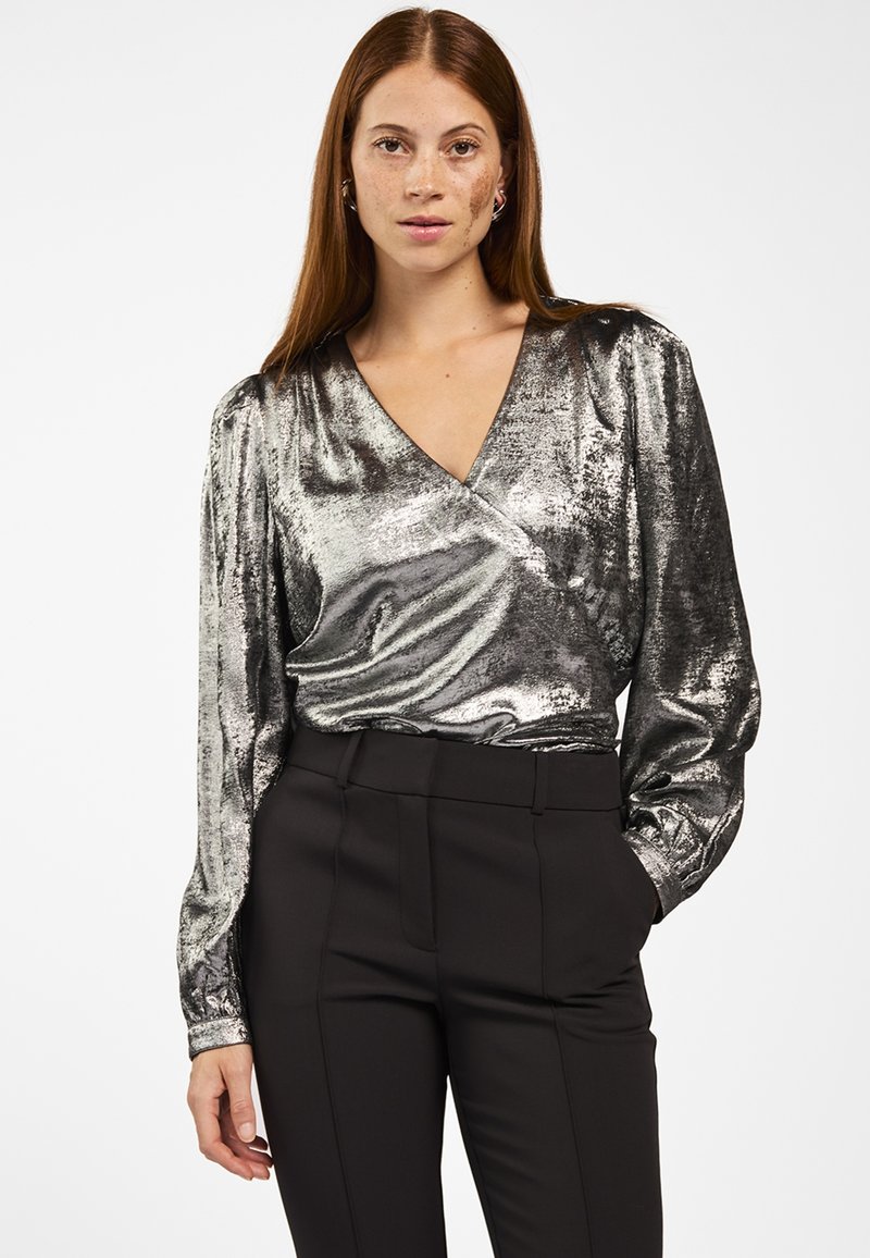 Vrouw met lang kastanjebruin haar, gekleed in een glanzende metallic zilveren wikkelblouse en hoge zwarte broek, staande met één hand in haar zak.