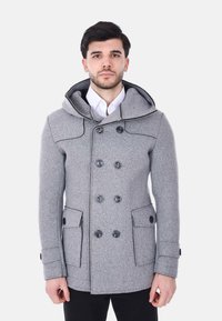 Uomo che indossa un cappotto doppiopetto di lana grigia con bottoni neri, cappuccio, tasche con patta, camicia bianca e pantaloni neri, in piedi davanti a uno sfondo bianco.