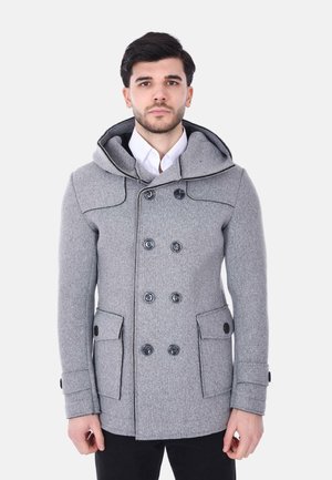 Uomo che indossa un cappotto doppiopetto di lana grigia con bottoni neri, cappuccio, tasche con patta, camicia bianca e pantaloni neri, in piedi davanti a uno sfondo bianco.