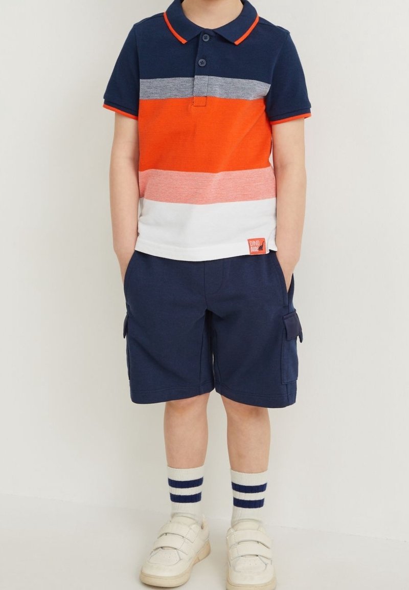 Enfant portant un polo à manches courtes rayé bleu, orange et blanc, un short cargo bleu marine, des baskets blanches et des chaussettes rayées.