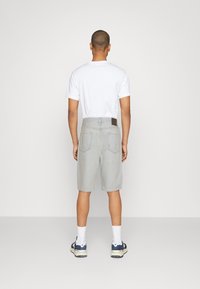 Pánské šedé džínové šortky s zadními kapsami, nošené s bílým tričkem a modrými teniskami. Hladká textura, délka ke kolenům a detail kožené visačky.