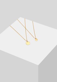 Goldfarbene Halsketten mit zwei runden Anhängern, einer größer und einer kleiner, die an dünnen Ketten vor einem minimalistischen Hintergrund hängen.