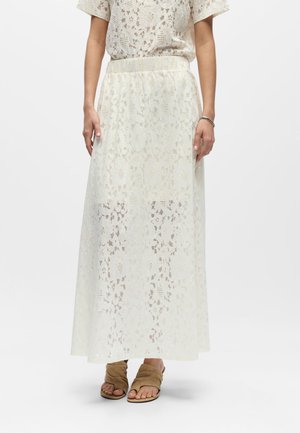 Jupe midi en dentelle blanche avec motif floral, portée avec un top en dentelle assorti et des sandales à talons ouvertes couleur beige sur un fond uni.