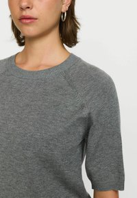 Vrouw in een grijs korte mouwen gebreid vest en zilveren ringoorbellen voor een eenvoudige witte achtergrond.