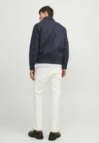 Tumedast sinine kangast jakk, millel on seisev krae ja elastne vöökoht, koos valgete denimipükste ja mustade seotud kingadega.