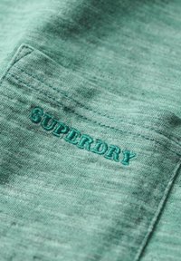Mintgrønt stof med en blød tekstur har et broderet "SUPERDRY" logo i matchende tråd. Synlige lomme detaljer forbedrer designet.
