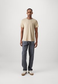 T-shirt beige a maniche corte con collo a contrasto, abbinato a jeans grigi e sneakers bianchi, in piedi davanti a uno sfondo neutro.