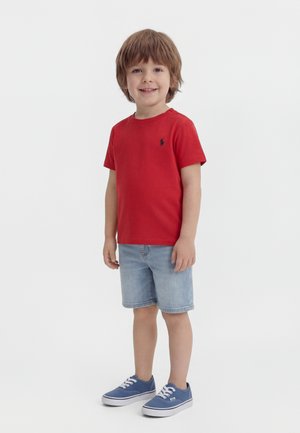 Jeune garçon debout, portant un t-shirt rouge à manches courtes, un short en denim bleu clair et des baskets bleues, souriant à la caméra sur un fond blanc.