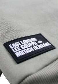 Tissu vert clair avec une surface texturée présentant une étiquette rectangulaire noire. L'étiquette affiche "EAST LONDON LEE COOPER BRITISH HERITAGE" en texte blanc.