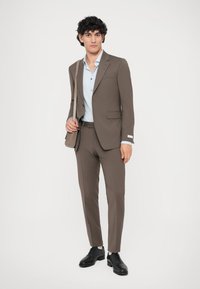Completo marrone con vestibilità su misura, giacca a un bottone e pantaloni slim. Camicia azzurro chiaro sottostante, abbinata a scarpe nere e una borsa a tracolla.