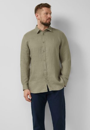 Mann mit Bart, der ein olivgrünes langärmeliges Hemd mit Knöpfen und dunkelblaue Hose trägt, steht vor einer hellgrauen Wand und schaut nach links.