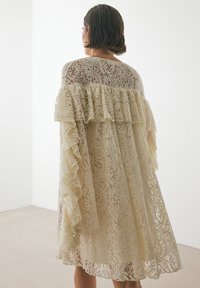 Spitzenkleid in zartem Beige mit gestaffelten Rüschen an den Schultern. Verfügt über filigrane florale Muster und eine lockere, fließende Silhouette.