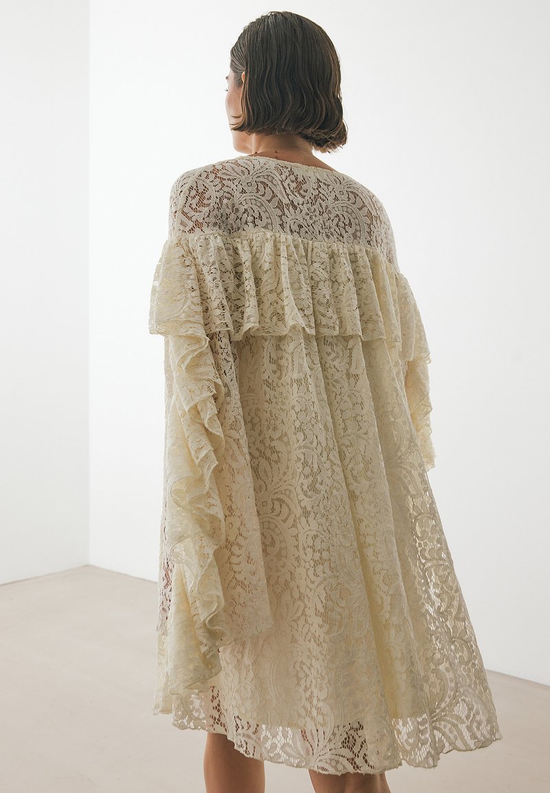 Spitzenkleid in zartem Beige mit gestaffelten Rüschen an den Schultern. Verfügt über filigrane florale Muster und eine lockere, fließende Silhouette.