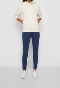 Kremowa bluza z kapturem z przodu i sznurkiem, połączona z granatowymi spodniami o wąskim kroju oraz białymi trampkami. Gładki materiał, casualowy design.