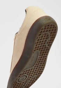 Zapato de ante y sintético en beige con una suela de goma oscura, que presenta un patrón de agarre texturizado y un detalle de logo embossado en el talón.