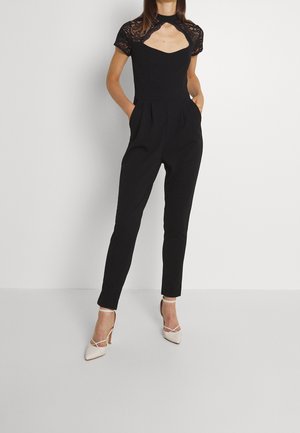 Combinaison noire avec manches courtes en dentelle, encolure avec ouverture en forme de goutte, et pantalon fuselé. Dotée de poches latérales et associée à des talons de couleur claire.