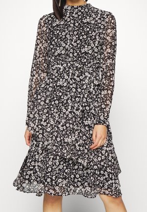 Czarna sukienka w białe kwiaty, z długimi, przezroczystymi rękawami, wysokim dekoltem, talią podkreśloną paskiem oraz falbaniastym, sięgającym kolan dołem, noszona przez stojącą osobę.