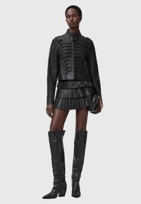 Zwarte leren cropped jack met decoratieve veters, gecombineerd met een geplooide zwarte minirok en zwarte leren laarzen tot aan de knie.