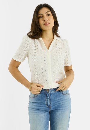 Mujer con blusa blanca de encaje de manga corta y jeans azules de cintura alta, posando con las manos cerca de la cintura frente a un fondo blanco.