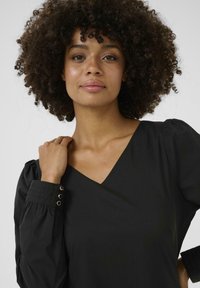 Blouse noire avec un décolleté en V, des manches bouffantes et des poignets texturés ornés de trois boutons dorés. Tissu lisse, silhouette ajustée.