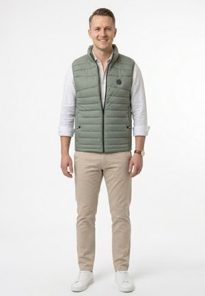 Hombre sonriente de pie, vistiendo un chaleco acolchado verde claro sobre una camisa blanca, pantalones beige, zapatillas blancas y un reloj de pulsera.