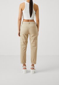 Kvinna med långt mörkt hår klädd i en vit kortärmad topp, beige byxor med uppvikta ben och vit bälte, samt vita plattformssandaletter, står framför en neutral bakgrund.