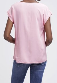 Blusa rosa con maniche corte e scollatura tondeggiante, realizzata in tessuto liscio. Abbinata a jeans blu, che mostrano un fit rilassato e una caduta fluida.