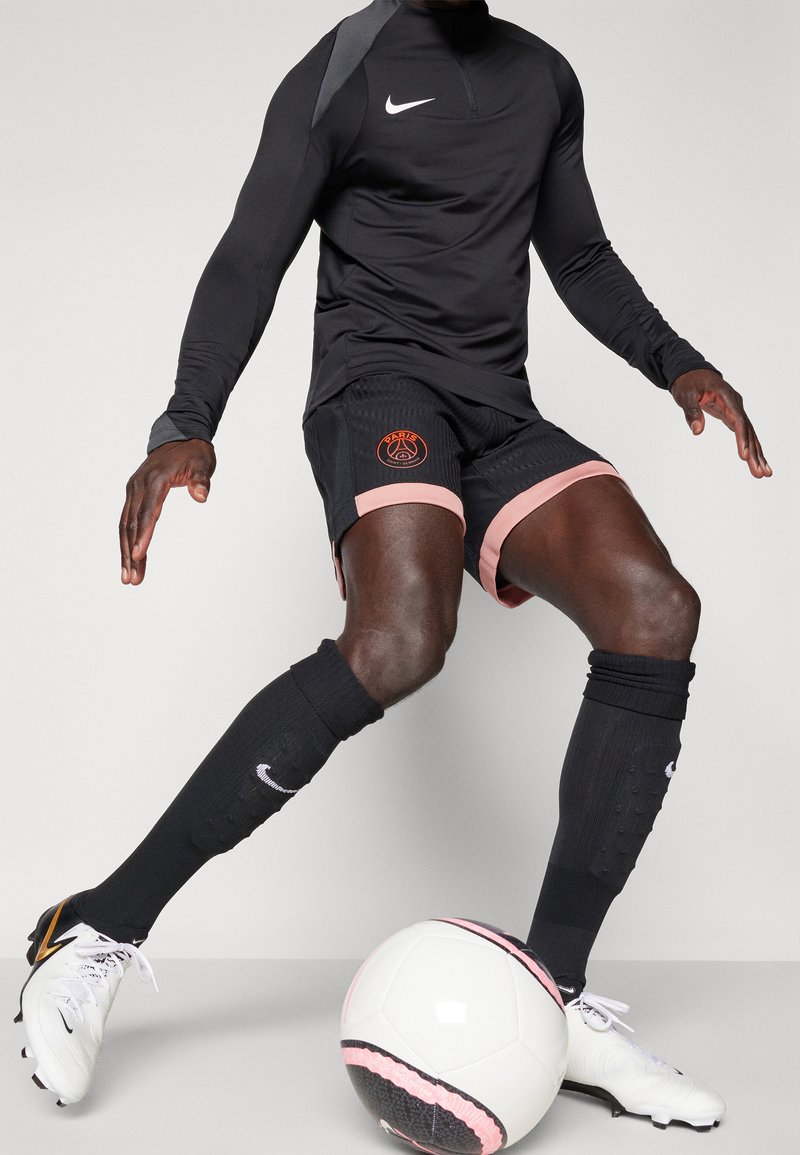 Schwarzes langärmeliges Nike-Shirt, schwarze Shorts mit rosa Akzenten und Paris-Logo, schwarze Socken und weiße Fußballschuhe. Weißer Fußball am Fuß.