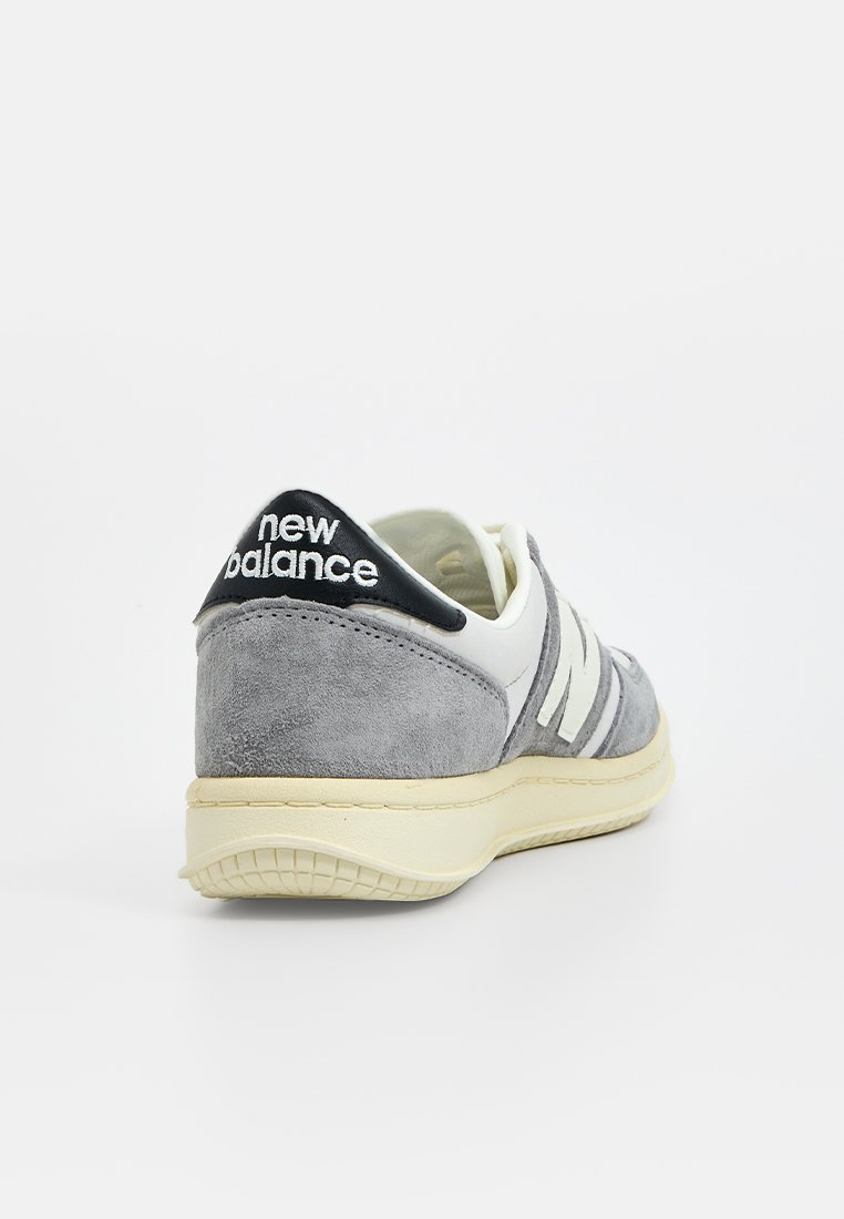 Zapatilla de ante gris con acentos en blanco, que presenta una pestaña negra en el talón con la marca "new balance" y una suela de goma texturizada.