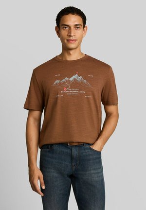 Mann trägt ein braunes T-Shirt mit Berggrafiken und Text "Explore Beyond Limits,", kombiniert mit blauer Jeans und braunem Gürtel, vor grauem Hintergrund.