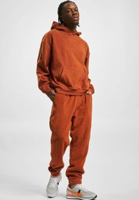 Oranžno flis s kapuco z kenguru žepom, v kombinaciji z ujemajočimi se trenirkami. Bele in sivo teniske z oranžnimi poudarki.