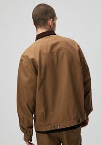 Mężczyzna z krótkimi włosami i kolczykiem w uchu, ubrany w brązową kurtkę z ciemnym kołnierzem, stojący tyłem do kamery na tle jednolitego tła.