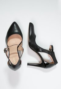 Chaussures à talons hauts noires avec bout pointu, bride réglable et finition brillante. Présentent un design ajouré sur les côtés et une texture en cuir.