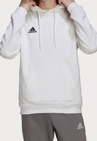 Bílá bavlněná mikina s nastavitelnými šňůrkami, přední klokanková kapsa a černé logo adidas na levém hrudi. Šedé tepláky viditelné dole.