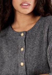 Frau in einer grauen gerippten Strickjacke mit drei goldenen Knöpfen, Lippen leicht geöffnet und lange dunkle Haare auf einer Seite sichtbar.