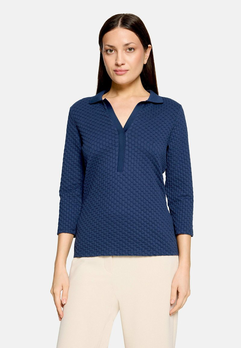 Femme aux longs cheveux foncés portant un polo bleu marine texturé à manches longues et un pantalon beige clair devant un fond uni.