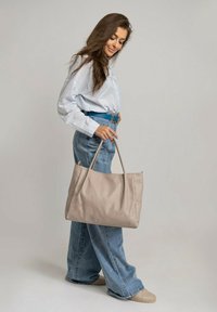 Beżowa skórzana torba na ramię z dwoma mocnymi uchwytami, elegancki fason i gładka tekstura, zestawiona z niebieskimi dżinsami z szerokimi nogawkami i białą koszulą.