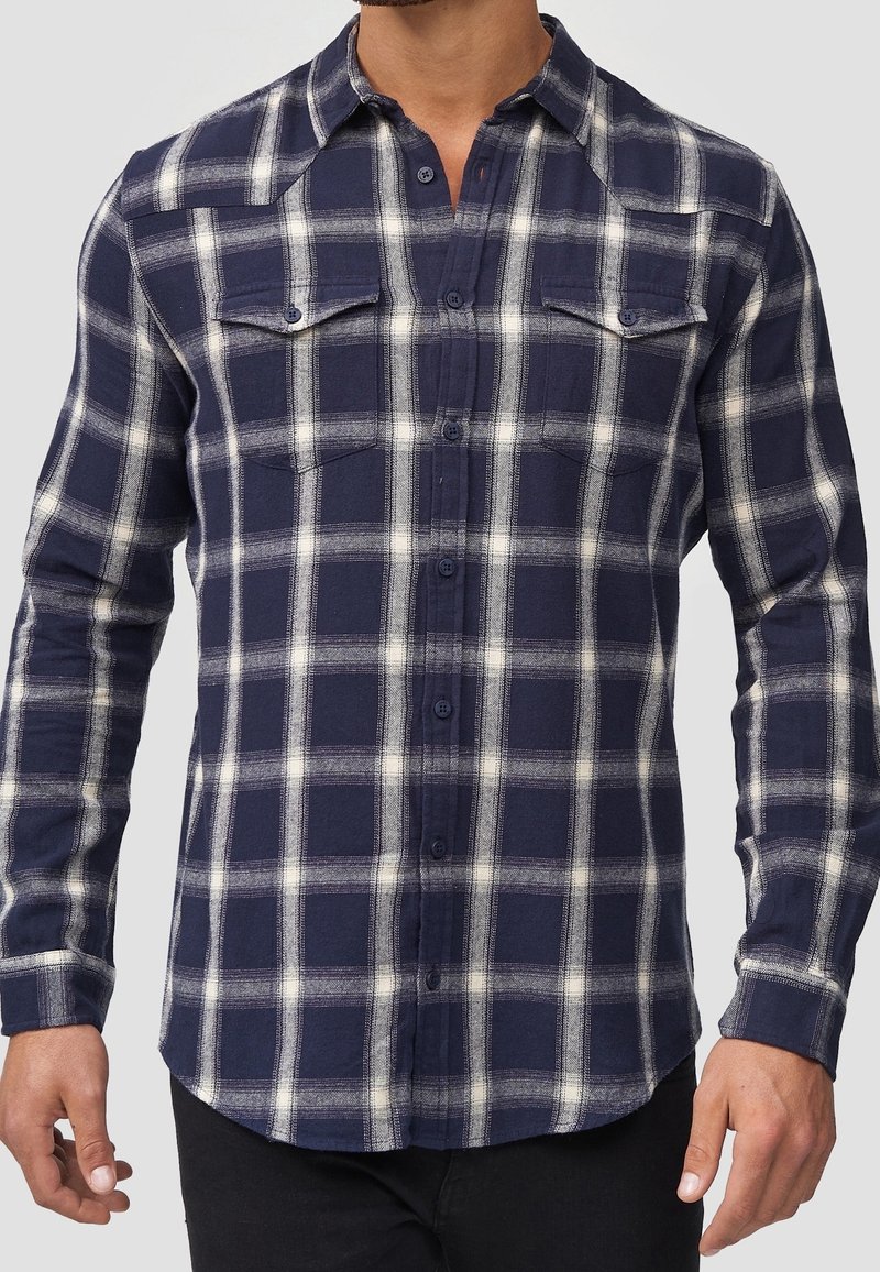 Homme portant une chemise à manches longues boutonnée à carreaux bleu marine et blanc avec deux poches poitrine, debout devant un fond neutre.