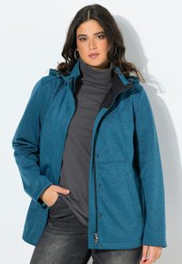 Nainen, joka käyttää turkoosia takkia tummanharmaan poolopaidan päällä, käsi taskussa, seisoo valkoista taustaa vasten.