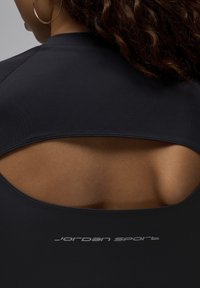 Czarny top sportowy z wycięciem na plecach, gładką teksturą i logo "Jordan Sport" w jasnoszarym kolorze na dolnej krawędzi.