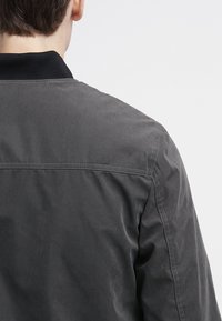 Šedá bomber bunda s žebrovaným černým límcem, hladkou strukturou a viditelnými švy podél ramen a zad.