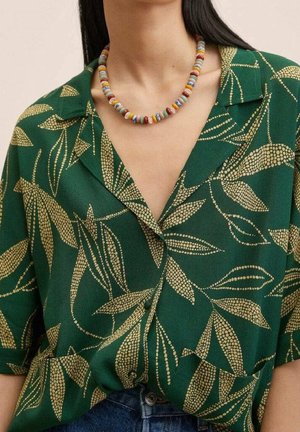 Femme portant une chemise verte avec des motifs de feuilles beiges et un collier de perles coloré, montrée du cou à la taille.