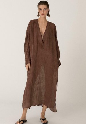 Frau trägt ein lockeres, langärmliges braunes strukturiertes Kleid mit tiefem V-Ausschnitt und schwarzen Sandalen, steht vor einem schlichten Hintergrund.