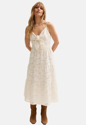 Frau trägt ein weißes Midikleid aus Lochstickerei mit dünnen Trägern und einer Schleife vorne, kombiniert mit braunen Wildleder-Stiefeln bis zur Mitte der Wade, steht vor einem schlichten Hintergrund.