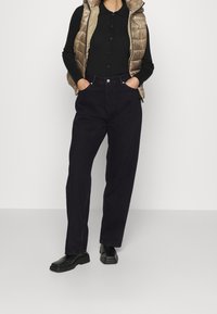 Mujer con blusa negra de manga larga abotonada, chaleco acolchado dorado, pantalones negros de cintura alta y zapatos negros de punta cuadrada, de pie con las manos en los bolsillos.