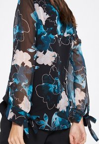 Durchsichtige schwarze Bluse mit blau-weißen Blumenmuster, mit Bändern an den Manschetten und einer lockeren Passform. Leichter Stoff sorgt für Textur.